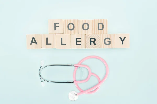 青色で隔離された聴診器の近くに食物アレルギーの文字を持つ木製のブロックのトップビュー — ストック写真