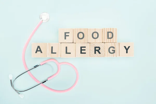 青色で分離された食物アレルギー碑文を持つ木製のブロックの近くのピンクの聴診器のトップビュー — ストック写真