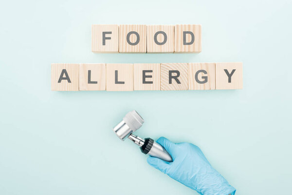 cropped view of doctor in latex glove holding dermatoscope near wooden blocks with words food allergy on blue