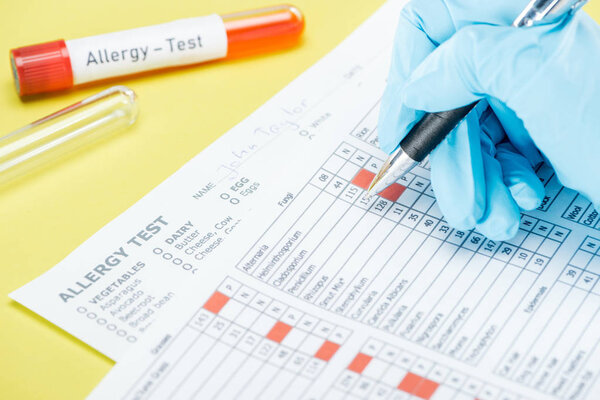 cropped view of woman in latex glove filling in papers near test tubes with allergy test lettering isolated on yellow