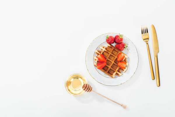 top view of waffle with strawberries on plate near bowl with honey and wooden dipper, and cutlery on white