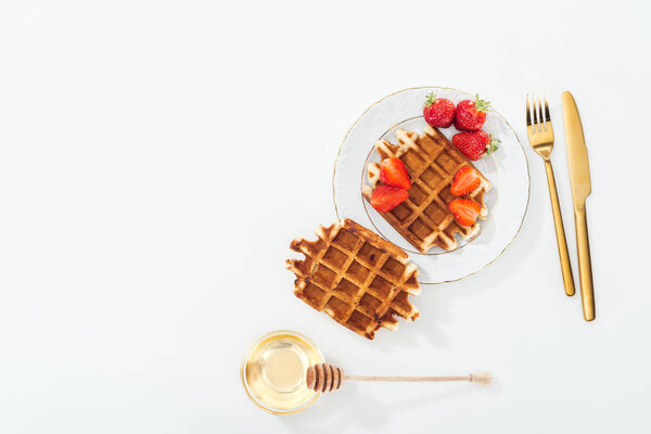 top view of waffles with strawberries on plate near cutlery, bowl with honey and wooden dipper on white