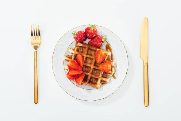top view of waffle and strawberries on plate and fork with knife near plate 
