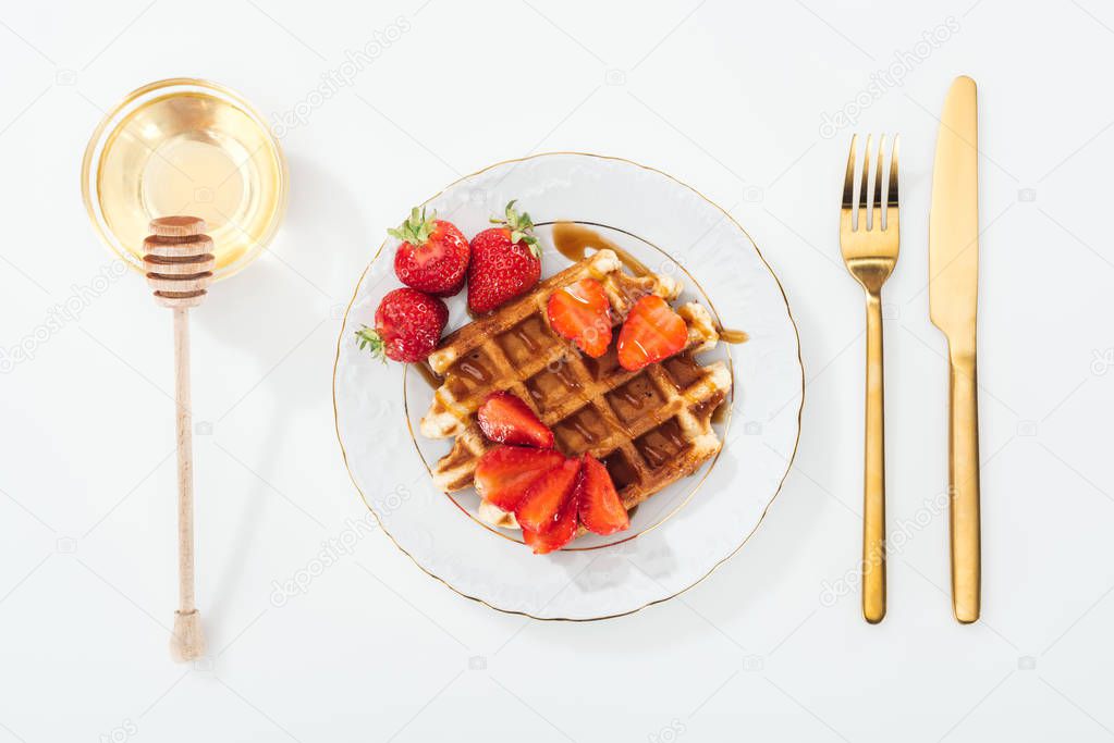 top view of waffle with strawberries on plate near cutlery, bowl with honey and wooden dipper 