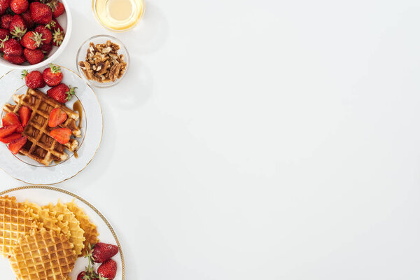 top view of tasty waffles and strawberries on plated near bowls with honey and nuts on white
