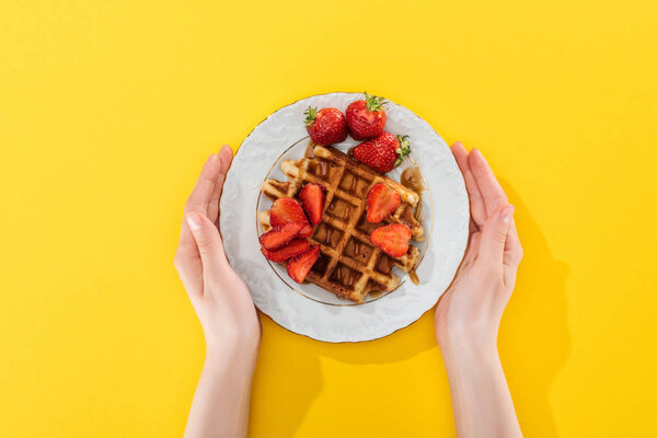cropped view of woman holding plate with waffle and strawberries on yellow