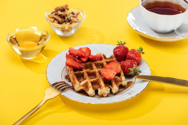 served breakfast with waffle and strawberries on plated near cutlery, honey, nuts and tea on yellow background