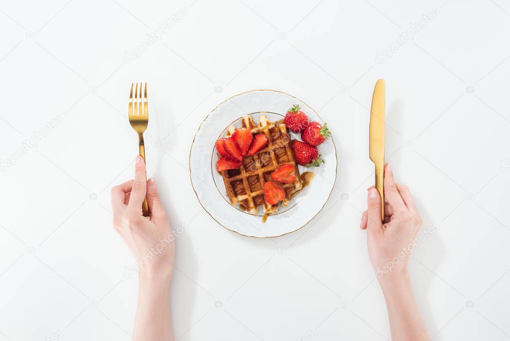cropped view of woman holding fork and knife near plate with waffle and strawberries on white