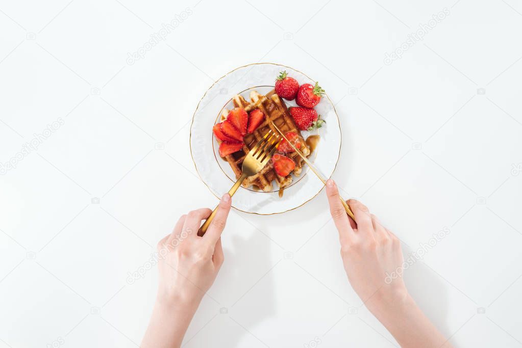 cropped view of woman cutting waffle with strawberries on plate on white
