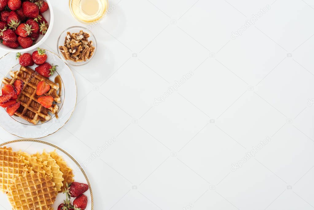 top view of tasty waffles and strawberries on plated near bowls with honey and nuts on white