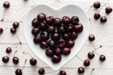 top view of fresh, sweet and ripe cherries covered with water drops on heart shaped plate  clipart