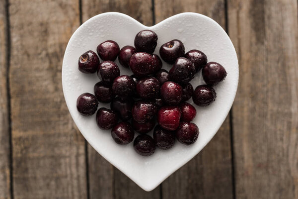 top view of red, fresh, whole and ripe cherries covered with droplets on plate on wooden surface 