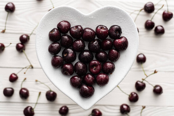 top view of fresh, sweet and ripe cherries covered with water drops on heart shaped plate 