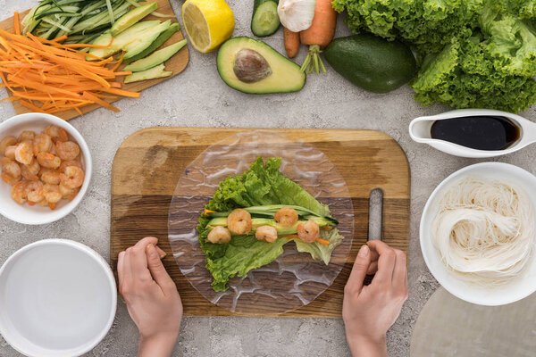 top view of woman making roll on cutting board with lettuce, shrimps and avocado 