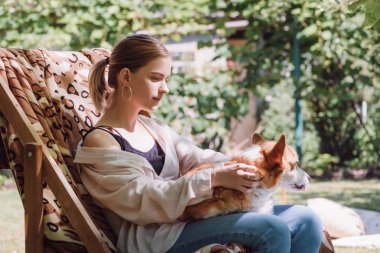 bahçede güverte sandalyede otururken dizlerde sarışın kız tutan Galce Corgi köpek