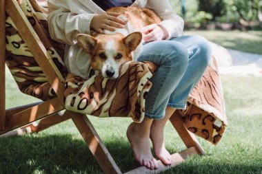 bahçede güverte koltukta otururken dizlerinin üzerinde çıplak ayakla Corgi köpek tutan yalınayak görünümü Kırpılmış Görünüm