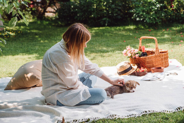 blonde girl sitting on white blanket in garden and having picnic at sunny day with cute puppy