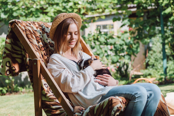 smiling blonde girl in straw hat holding puppy while sitting in deck chair in garden