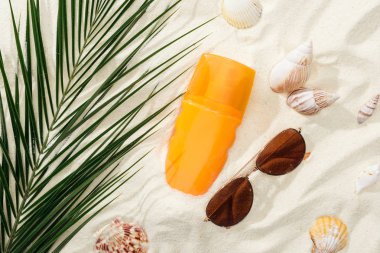 orange bottle of sunscreen on sand with seashells, green palm leaf and stylish sunglasses clipart