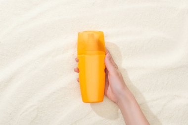 cropped view of woman holding orange sunscreen above golden sand clipart