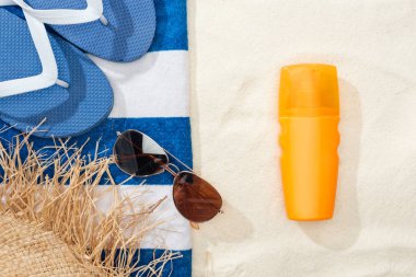 Güneş kremi, saman şapka, Flip flop, güneş gözlüğü ve altın kum üzerinde çizgili mavi ve beyaz havlu üst görünümü