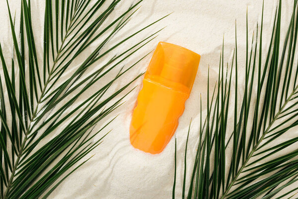 orange bottle of sunscreen on sand with green palm leaves
