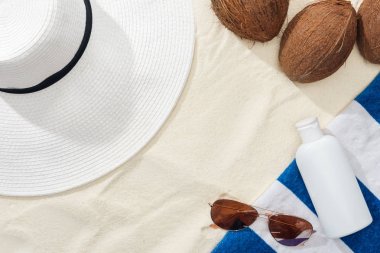 top view of sunglasses, striped towel, coconuts and white straw hat near white lotion on sand clipart