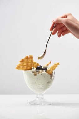 cropped view of woman holding spoon near delicious pistachio ice cream with waffles and blueberries isolated on grey clipart