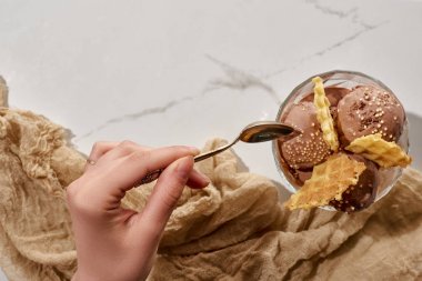 cropped view of woman holding soon near delicious chocolate ice cream in bowl with waffles and brown cloth on marble grey background clipart