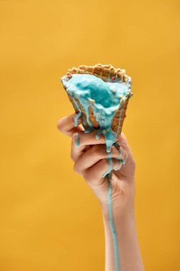 cropped view of woman holding melted blue ice cream in crispy waffle cone isolated on yellow clipart