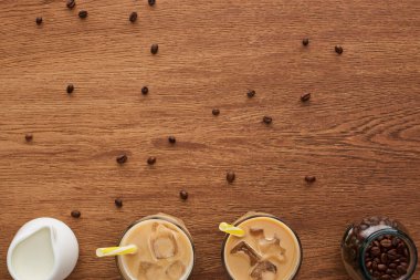 top view of coffee in glasses with ice and straws, milk and coffee grains on wooden table with copy space clipart