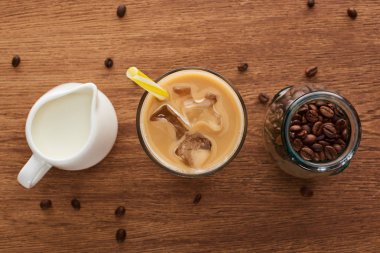 top view of ice coffee with straw near milk in jug and coffee grains in jar on wooden table clipart