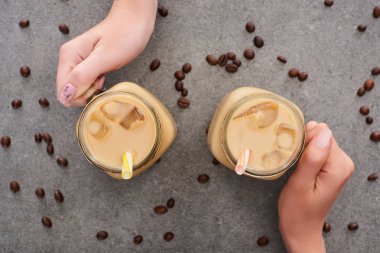  cropped view of women holding glass jars with ice coffee and coffee grains on grey background clipart
