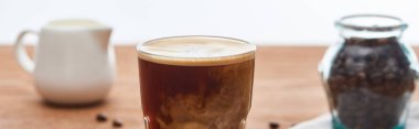 Beyaz, panoramik çekim üzerinde izole ahşap masa üzerinde süt sürahi ve kahve taneleri yakın cam süt ile buz kahve seçici odak