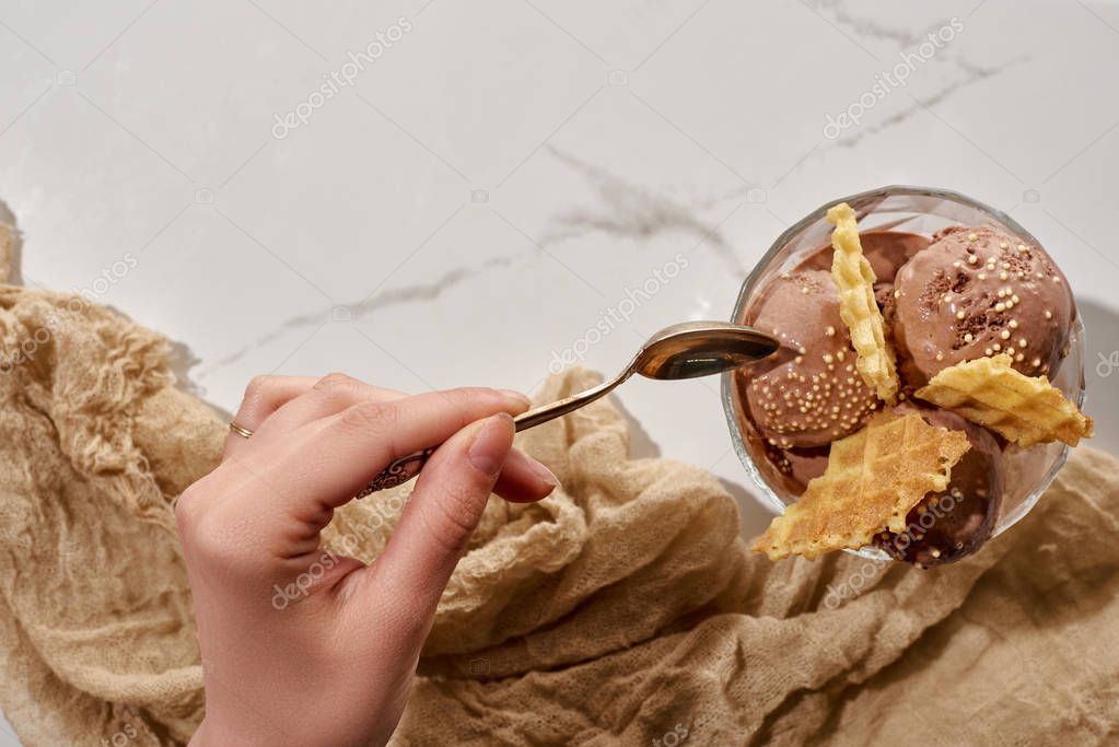 Cropped view of woman holding soon near delicious chocolate ice cream in bowl with waffles and brown cloth on marble grey background