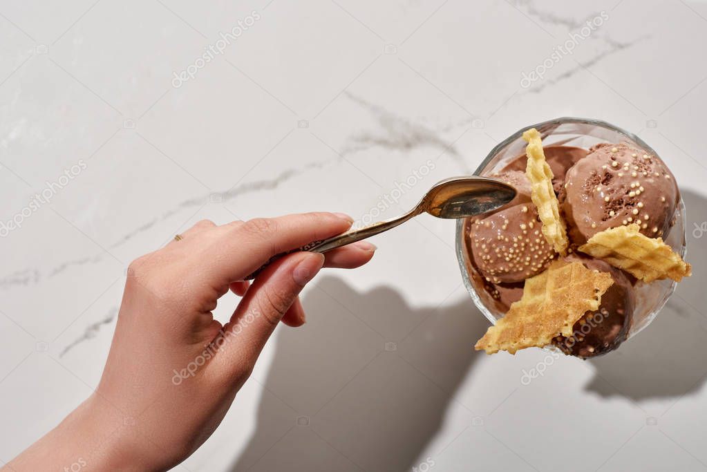 Cropped view of woman holding soon near delicious chocolate ice cream in bowl with waffles on marble background