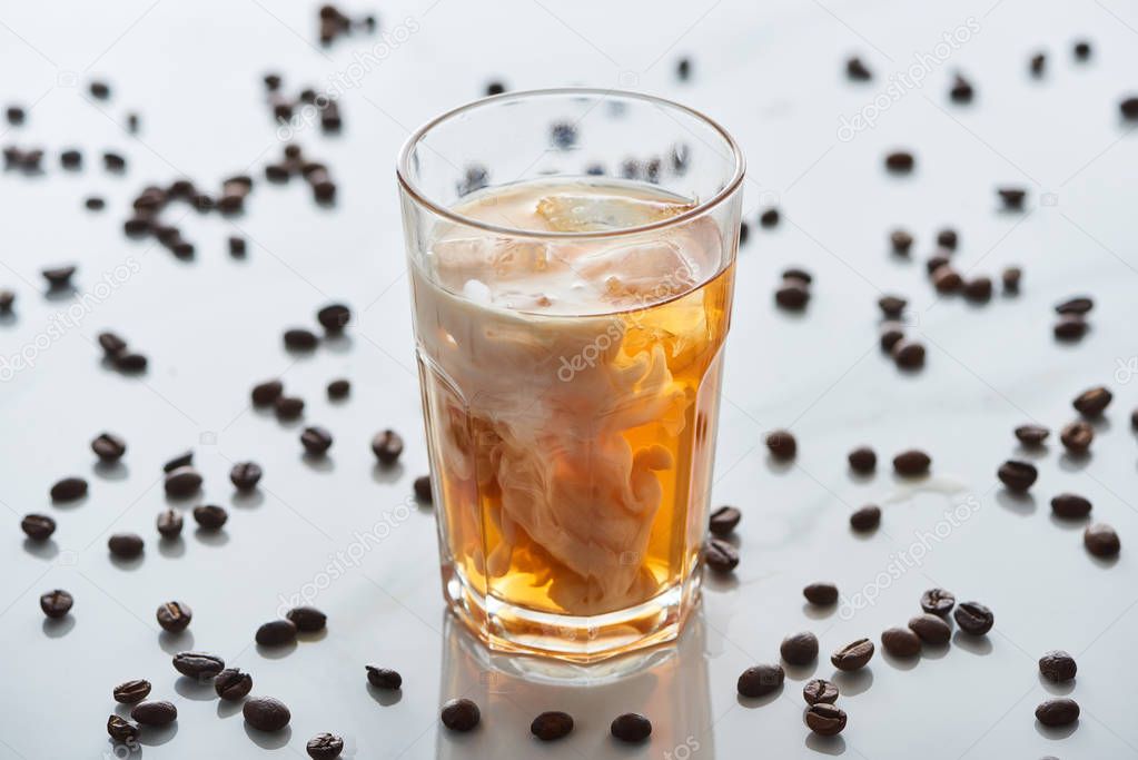 selective focus of ice coffee mixing with milk in glass near scattered coffee grains on grey background