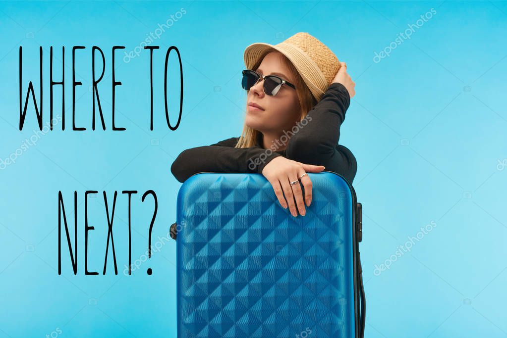 blonde girl in sunglasses and straw hat near blue suitcase isolated on blue with where to next question