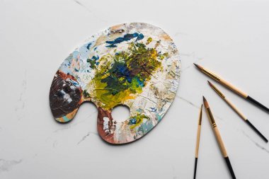 top view of colorful palette with oil paints near paintbrushes on marble white surface