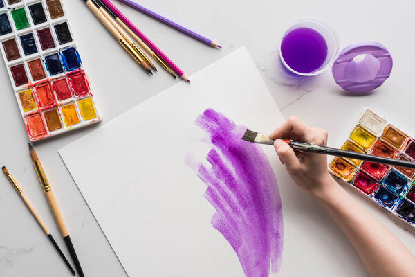 обрезанный вид художника, рисующего фиолетовые акварельные мазки на белой бумаге на мраморной белой поверхности
