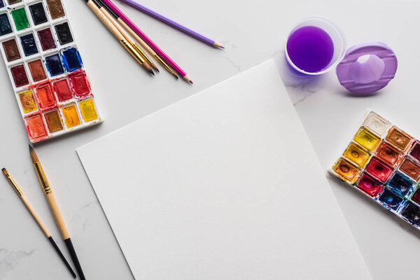вид сверху на пустую белую бумагу возле кистей и акварельных красок на мраморной белой поверхности
