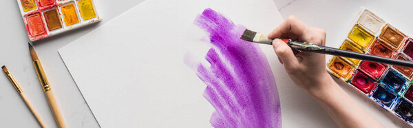 обрезанный вид художника, рисующий фиолетовые акварельные мазки на белой бумаге на мраморной белой поверхности, панорамный снимок
