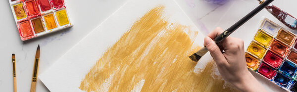 обрезанный вид художника, рисующего желтые акварельные мазки на белой бумаге на мраморной белой поверхности, панорамный снимок
