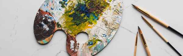 top view of colorful palette with oil paints near paintbrushes on marble white surface, panoramic shot
