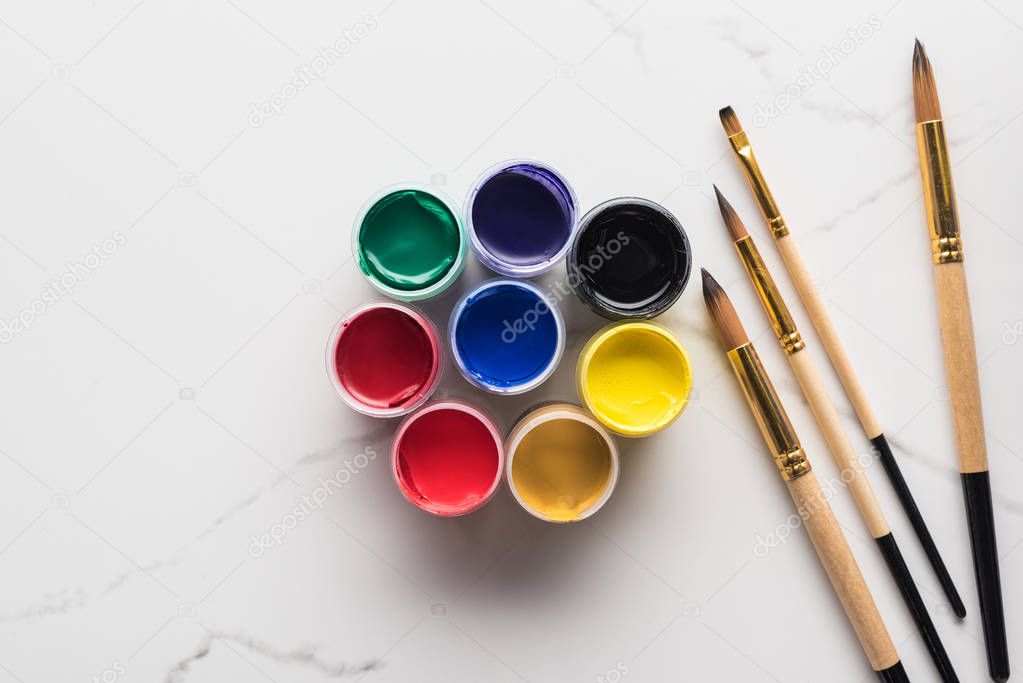 top view of multicolored gouache paints and paintbrushes on marble white surface
