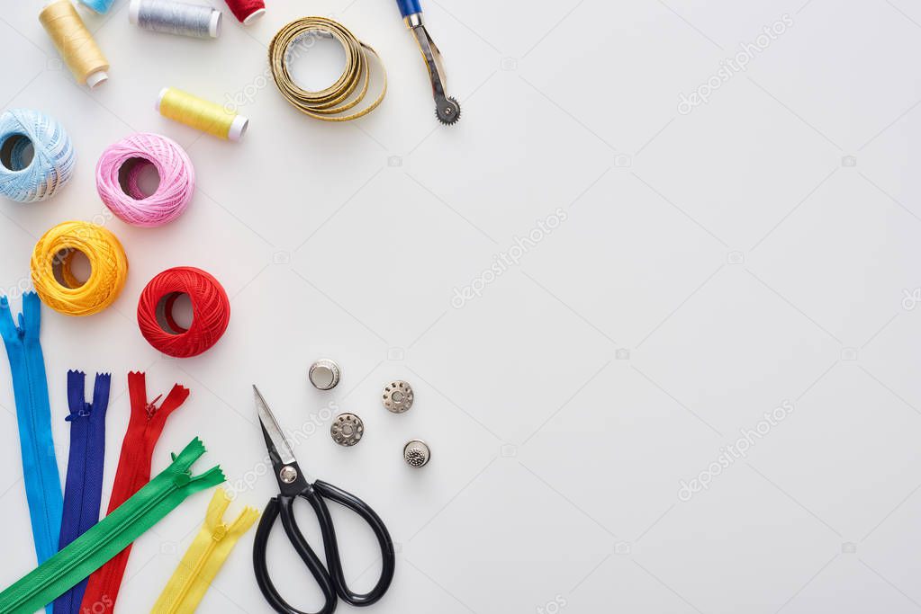 top view of zippers, scissors, thimbles, threads, knitting yarn balls, bobbins, tracing wheel, measuring tape on white background 