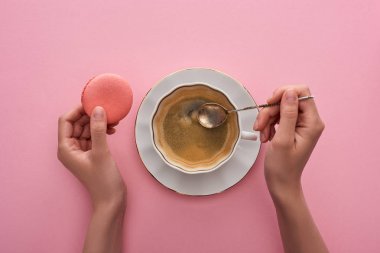 partial view of woman holding spoon near coffee in cup and delicious French macaroon on pink background clipart