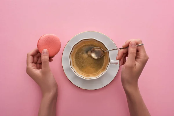 カップのコーヒーの近くにスプーンを保持し ピンクの背景においしいフランスのマカロンを保持している女性の部分的なビュー — ストック写真