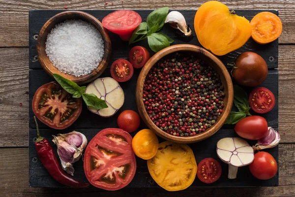 top view of bowls with pepper and salt near chilli pepper, spinach, sliced tomatoes and garlic on black tray on wooden table 