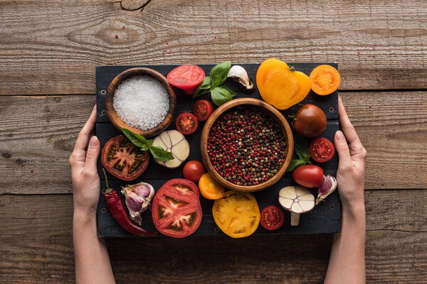 cropped view of woman holding black tray with pepper and salt in bowls, chilli pepper, sliced tomatoes and garlic on wooden table 